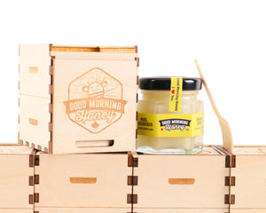  A small wooden crate with the logo "Good Morning Honey" engraved on it is placed on a stack of similar crates. Next to the crate is a small glass jar of honey with a yellow label that reads "Good Morning Honey" and "Miel Biologique." A wooden honey dipper is leaning against the jar.