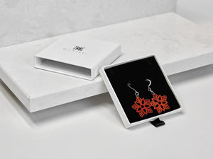 A pair of red flower-shaped earrings with intricate cut-out patterns, displayed in a white box labeled "OHTSII."